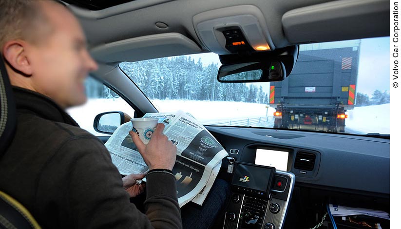 Inside view of a vehicle that is part of a platoon. The photo shows the driver of the vehicle, as well as the Lead Truck in the platoon through the front windshield. The scene outside shows a snowy day, the narrow highway lined with evergreen trees. The driver is able to enjoy a cup of coffee and read the paper without having to steer the car; he is smiling.