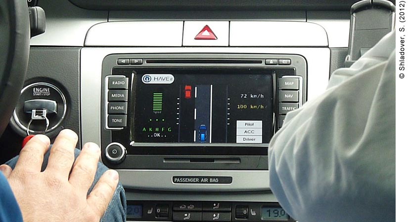 View of a digital display on a car dashboard. A driver and a passenger are in the front seat of the car; only the driver’s knee (with hand on it) and the passenger’s elbow can be seen. Between the driver and the passenger is the dashboard display for Volkswagen’s Temporary Auto Pilot. The Temporary Auto Pilot has the HAVEit logo on the top left hand corner of the screen. Below the HAVEit logo are green horizontal digital bars on the screen, with the letters “A,” “K,” “H,” “F,” and “G” written below. Below the green letters is the word “OK in white. In the middle of the screen is a digital representation of two cars: a red one in the left lane ahead of a blue car in the right lane. To the right of the cars it says “72 km/h” in white and “100 km/h” in yellow. Below the speeds are three white bars: the top one says “Pilot”; the middle one says “ACC”; the bottom one says “Driver.” Surrounding the Temporary Auto Pilot screen are the regular features of the dashboard of any modern car: a CD player is above the screen, as well as the indicator light for emergency lights; buttons for “Radio,” “Media,” “Phone,” “Tone” and the Volume button for the stereo are to the left of the screen; buttons labeled “Map,” “Nav,” and “Traffic” are to the right of the screen (the passenger’s elbow is blocking the fourth button). 