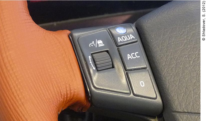 Close-up photo of the Automated Queue Assistance (AQuA) control panel buttons on a steering wheel. The control panel is made up of three buttons and a dial. The dial (on the left) shows the Adaptive Cruise Control (ACC) dashboard indicator symbol. To the right of the dial are the buttons that indicate the three levels of driver control: “AQuA,” “ACC”, and “O”. AQuA is designed for highly-automated driving in congested traffic.