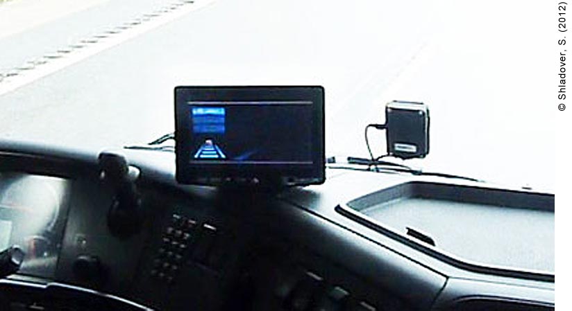 Close-up view of the Automated Queue Assistance (AQuA) driver interface screen. The device is mounted on the dashboard, much like a GPS device would be. A sensor mounted on the inside the front window is attached to the device. The screen is too blurry to read, but most AQuA devices display the three levels of driver control: AQuA (highly-automated), ACC (adaptive cruise control; partial automation), and Manual (not automated). It appears that the AQuA level is activated because the highest indicator is lit.