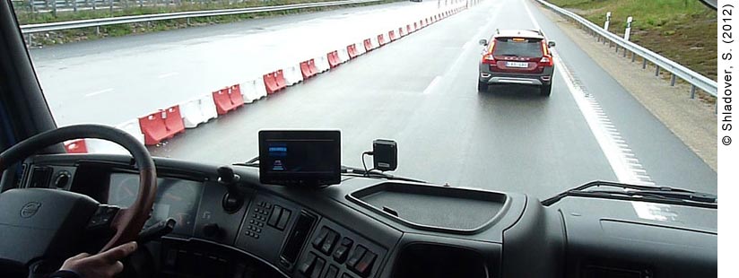 Photo inside the cab of a truck. The driver is cropped out of the photo, but his hands are visible on the steering wheel. On the dashboard is an Automated Queue Assistance (AQuA) device, indicating that the truck is using ACC (adaptive cruise control). A sensor attached to the AQuA device is attached inside the front windshield. The view through the windshield is of a four-lane highway, divided in the center by red and white traffic barriers. A red Volvo can be seen driving right in front of the truck through the windshield.