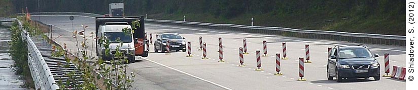 Daytime view of a four-lane highway, with pylons diverting traffic to create a new lane. Two oncoming cars are driving through the pylon-created lane. A truck in the lane to the left is driving closely to the pylon lane but has not entered it yet. Aside from the cars and the truck, there are no other cars on the highway.