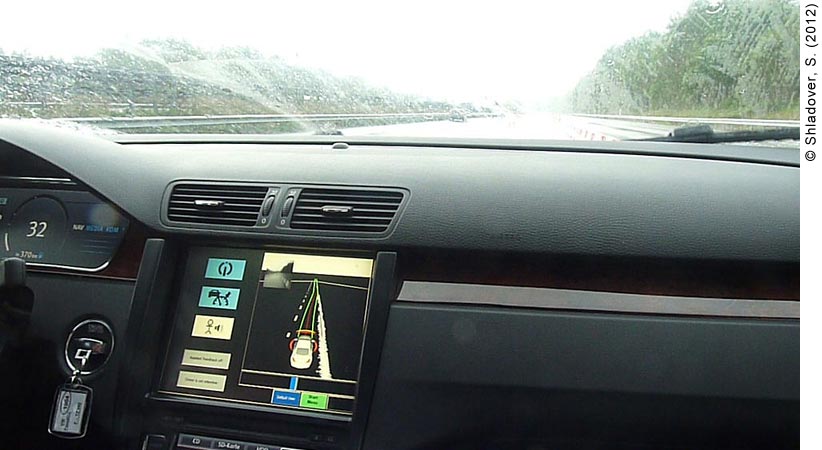 . Photo of a car dashboard equipped with a Roadworks Assistant driver-vehicle interface (DVI). Rain is falling, making it difficult to see clearly through the front windshield; it looks like the car is driving on a tree-lined highway. The DVI screen is located on the dashboard just below the heating/AC vents to the left of the steering wheel.  The screen on the device shows a digital representation of a car on a highway. Red and green lines are emanating from the front of the digital car on to the digital highway. Red and yellow lights can be seen perpendicular to the digital car in the front; red lights are surrounding the digital car on the sides. To the left of the screen are five touch-screen buttons. The top button shows the HAVEit logo in a blue box (highly-automated mode); below that is another blue box with the dashboard icon that indicates the semi-automated mode. Below the semi-automated icon is a stick figure with a steering wheel, indicating the Driver Assisted mode; the icon is white, indicating that the car is being driven in this mode.