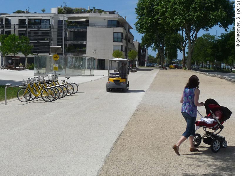 . Daytime view of a pedestrian walkway in La Rochelle, France. A wide, paved pedestrian path is in the center of the photo; a yellow CyberCar is approaching on the path. To the left of the CyberCar is a public bike-sharing rack filled with yellow bikes. Behind the bike rack is row of four-story apartment buildings; the closest one has a garden on the roof. To the right of the pedestrian path is a tree-lined street; a woman pushing a baby carriage is walking on the path toward the street.