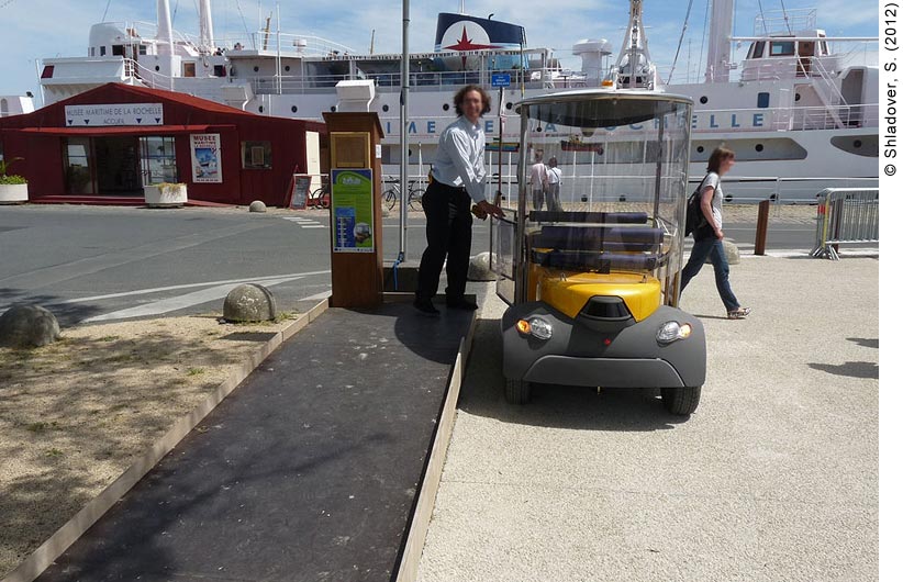 Daytime photo of a man opening the door of a CyberCar at a CityMobil pick-up point at the port in La Rochelle, France. The CyberCar is yellow and gray, with glass enclosing the passenger area. Four passengers (two facing forward, two facing back) can fit in this CyberCar, which currently has no passengers. The man is standing in the parking lot of the Maritime Museum (Musée Maritime) La Rochelle. In the background is a small red wooden building with “Musée Maritime de La Rochelle” written on it. Behind the building is a docked white cruise ship. A woman is walking away from the museum to the right. A couple can be seen looking at the ship.