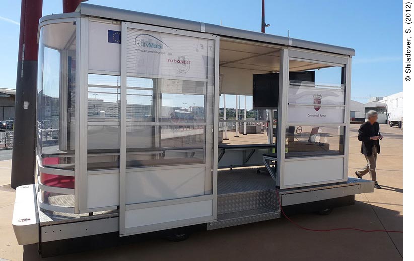 Daytime view of an empty 22-passenger CyberCar. The CyberCar looks similar to a small shuttle bus. The CyberCar is enclosed in glass, with benches lining the walls for seating. A sliding glass door is open, revealing a metal step to allow passengers to get on. Large silver bumpers extend out the front and rear. Inside a large flat-screen TV is mounted on the right rear corner. A man is walking behind the CyberCar to the right.