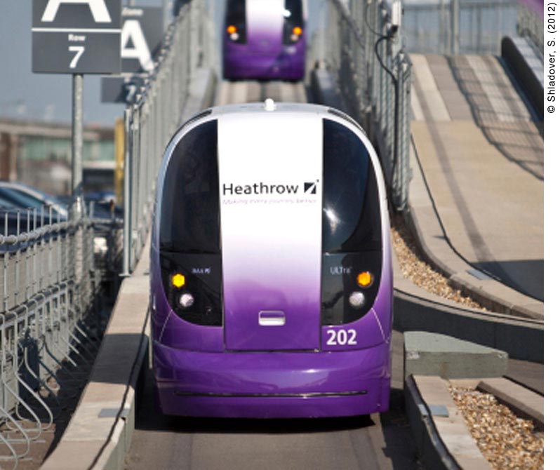 Two personal rapid transit vehicles (PRTs) approaching a station on a track at Heathrow Airport. The PRTs (known as pods at Heathrow) are both purple and white, and are approaching on a single-lane paved track. To the left of the track is an airport parking lot: cars are parked in the lot and location signs are posted indicating the Zone and Row that the cars are parked in. On the right is another pod track (presumably for pods travelling in the opposite direction), which is currently empty. A metallic overhang reflects the colors on the on-coming pods at the top of the photo. 