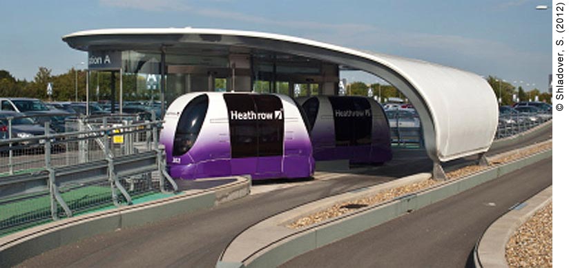 Two personal rapid transit vehicles (PRTs) parked at Station A in a parking lot at Heathrow Airport. The PRTs (known as pods at Heathrow) are purple and white and have the Heathrow logo written on the side windows. The pods are small, only carrying six passengers at a time. The pod station is a small glass enclosure; a sign reading “Station A” is partly visible around the top left hand side of the enclosure. Behind the station is long-term airport parking lot.