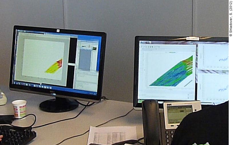 Two computer monitors displaying real-time plots of distance-versus-time diagrams. The monitor on the left shows a graph with a shock wave pattern of yellow bleeding in to red that goes up and to the right. The monitor on the left shows blue and green streaks in a shock wave pattern, also going up and to the right. The upper right hand corner of the second pattern shows white gaps in a field of green.