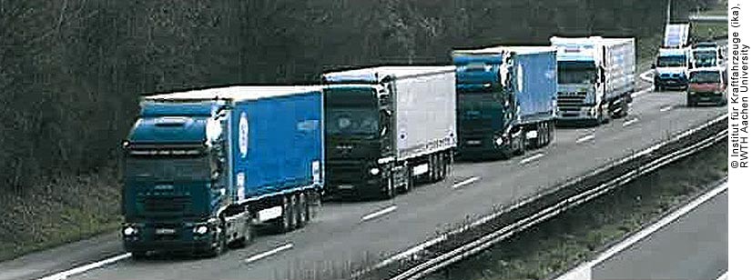 An oncoming KONVOI platoon of four trucks driving on a highway in the right lane. The highway is lined with trees that do not have many leaves; it looks like it is the end of fall. The highway is divided by a narrow center median with grass and bare-looking shrubs growing there. A red van is behind the platoon in the left lane; directly behind the platoon is a white van.