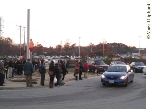 Early morning photo of a street corner. A Park-and-Ride lot surrounded by trees can be seen in the background; the sun is rising behind the trees. On the left of the photo about 10 people are lining up near a stop sign on the sidewalk. A blue sedan is approaching the line of people to the right of the curb with its headlights on.