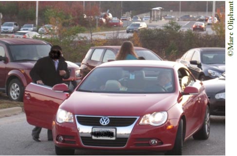 Two cars are lined up to pick up passengers at a Park-and-Ride lot. The car in front is red and a man is getting in to the passenger side. A woman is walking toward the second car in line, which is black. A parking lot surrounded by autumn trees can be seen in the background.