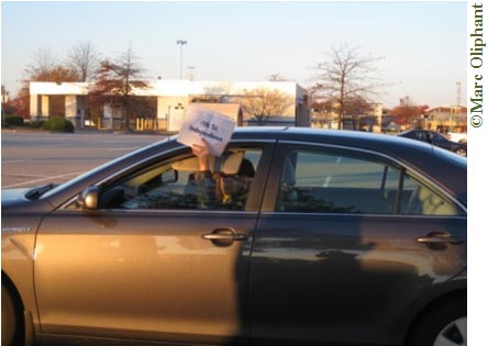 A daytime photo of the driver’s side of a gray four-door car. The sun is shining into the car. The car is parked in an almost empty parking lot. In the background past the parking lot is a white building and bare trees. The driver of the car is holding a sign out of his or her window (the writing is too small to see in the photo).