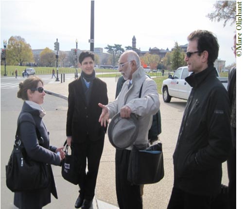 A woman and three men are standing together in a parking lot. It is daytime, and the parking lot is surrounded by trees and grass; a red brick building can be seen in the distance. The woman is on the left, and she is talking to the man who is second from the right; the other two men are listening. It is a bright day: the woman and the man on the far right are both wearing sunglasses.