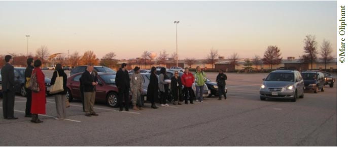 A daytime photo of a parking lot. Thirteen people are lined up on the left side of the photo in front of some parked cars. Beyond the parking lot are trees whose leaves have changed to fall colors. It appears to be cold: the people are all wearing coats, jackets, and sweat shirts. Three cars are driving up to the line of people from the right side of the photo, the first one with its headlights on.