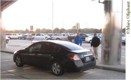 A black car is parked at the curb of a median and two men are standing next to the car on the median. Two concrete supports of what appears to be an overpass are on the median. Beyond the median is a parking lot filled with cars. Sunlight is streaming in: the sun is either rising or setting. Trees can be seen in the distance.