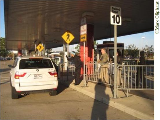 . A white SUV is parked at the curb of a median at a Park-and-Ride lot.  The median is lined with various street signs: two yellow crosswalk signs and a 10-mile an hour speed limit sign. Waist-high metal fences line the median, indicating where people can line up to wait for rides. Three people are lined up behind the fence. The person at the front of the line is trying to get the attention of the driver of the SUV by holding up his finger. Above the median is a weather shelter that covers the median and extends over the curb to cover cars as well.