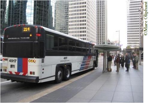 A white bus with red and blue stripes approaches a bus shelter on a downtown city street. A line of about four people has formed at the shelter. White skyscrapers can be seen on both sides of the street; a glass skyscraper across the street reflects the scene.