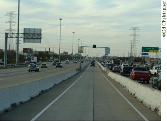 Photo of a highway on a cloudy day. Traffic is oncoming on the left side and is outgoing on the right. There is little traffic on the left; the traffic on the right is very congested. Between the oncoming and outgoing traffic is a center limited-access lane. Traffic in this lane is outgoing, but is less congested than the right lane. Above the limited-access lane is a traffic light and signs on either side (the signs are too blurry to read). There are billboards and exit signs on both sides of the highway. Light posts can be seen on the left side of the photo, but the lights do not appear to be on. Two large telephone towers are on either side of the highway, with telephone lines crisscrossing the skyline.