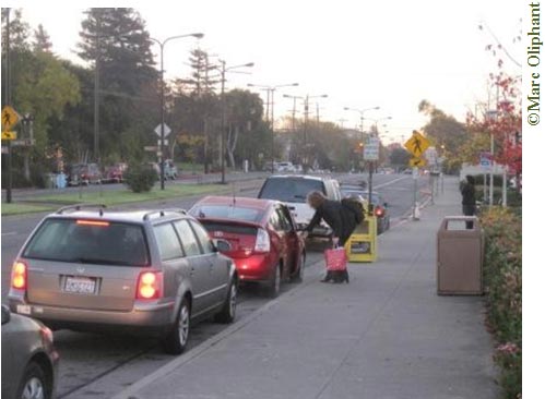 Early morning on a fall day. A line of cars are parked along the curb of a tree-lined street. The cars are all facing away from the camera on the right side of the street. Aside from the queuing cars, there is no traffic and there are no other parked cars on this side of the street. A woman is standing on the curb, opening the passenger-side door of the third car in line. The woman is wearing a backpack and is carrying a red bag. Just past the woman there is a brown trash can, a yellow newspaper dispenser, a parking sign, and a crosswalk sign. There is a grassy median in the middle of the street; tall street lights line the median but they are not on. Cars are parked on the other side of the street.