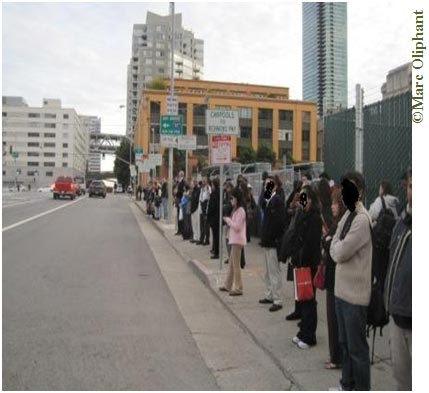 A daytime photo of a wide downtown city street. A long line of people is standing on the curb on the right side of the photo. Street signs line the curb; they are blurry, but appear to be parking signs, carpool signs, and signs that direct drivers to highway on-ramps. Behind the queue of people a chain link fence lines the sidewalk. A few cars are driving down the street, approaching a green traffic light. Beyond the traffic light there are buildings on both sides of the street, and a pedestrian overpass connects two of them.