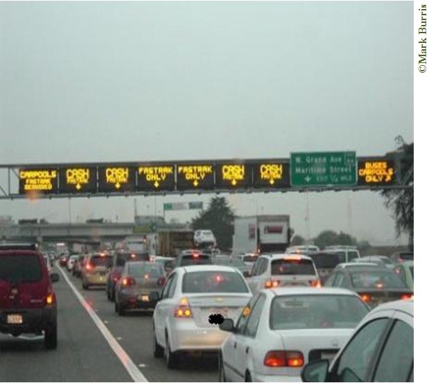 Outgoing highway traffic on a cloudy day. Traffic is bumper-to-bumper as it approaches digital tolling signs indicating which lanes to be in. There appear to be eight lanes of traffic. The digital signs indicate which lanes are for carpools, cash, FasTrak (an electronic toll collection system used in California), and buses. Carpools are directed to the two outer lanes; people paying with cash are directed into the second, third, sixth, and seventh lanes. The two middle lanes are for FasTrak-only drivers (FasTrak can also be used in the second thru eighth lanes). The far right lane is for buses and carpools only. Just before the digital tolling signs there is an exit sign on the right. An overpass with its own exit signs can be seen in the distance.