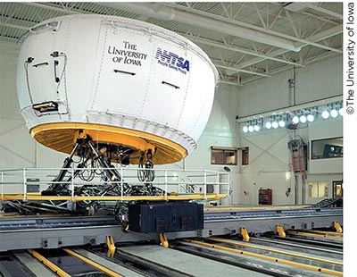 A photo of a driving simulator in a laboratory. The movable simulator is raised up on hydraulic legs and is branded with the logos of The University of Iowa and NHTSA.