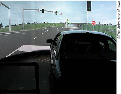 A photo of a sedan in front of a large wrap-around screen displaying a simulation of an approaching intersection.