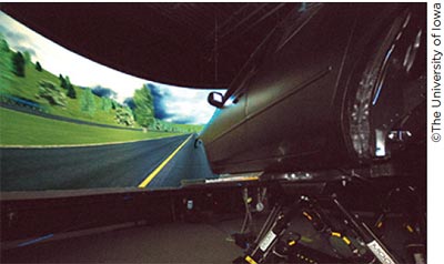 A view of a sedan mounted on a movable platform in front of a large wrap-around screen that is displaying a simulated highway.