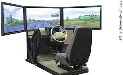 A photo of a driving seat, steering wheel, dashboard, and pedals in front of three monitors displaying a wrap-around simulation of a highway.