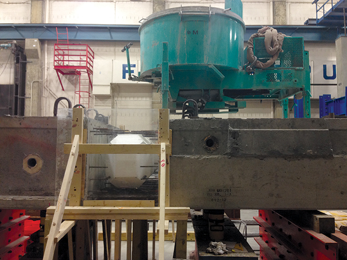 A photo of a laboratory. A view from the top of a ladder looking at a large green piece of machinery used to cast the beam splice connection.