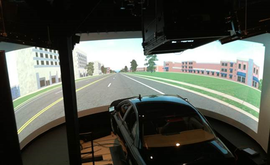 Title: Figure 4-2: Photograph. Driving simulator used for the study. - Description: The photo shows the driving simulator used for the study. It consists of the body of a car, which sits in front of a wraparound screen. The car sits on a base that moves to simulate the car's motion. The screen shows a simulated roadway environment. In the car's direction of travel, there are two lanes separated by a white dashed line. The opposite side of the roadway also has two lanes, separated by white dashed lines. The roadway is divided by two solid yellow lines. There are buildings, trees, and grassy areas on each side of the road.