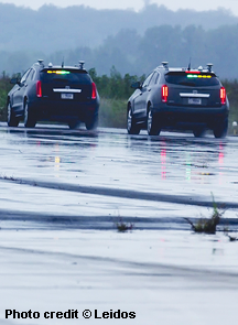 Two sport utility vehicles are traveling on a wet roadway. One vehicle is in front of the other. At the top of each vehicle’s back windshield are lights flashing green and yellow. Antennas are located on top of the vehicles, above the lights on the back windshields. The vehicles are simulating a platoon using cooperative adaptive cruise control, which combines sensors and vehicle-to-vehicle communication to enable vehicles to adjust their speed to the preceding vehicle in their lane. In the distance beyond the roadway is a wooded area. 