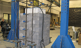 The photograph depicts a vertical column, square in cross-section, consisting of 10 tiers of rectangular masonry blocks. The column stands on the floor of a large industrial room. Vertical steel beams, painted blue, are seen to the left and right of the masonry column. A man is seen standing behind a low shelf of instruments at the left. A large concrete block connected to a cylindrical vertical shaft is pressing down on the top of the masonry column. Cables and wires are attached at various positions on the masonry column.