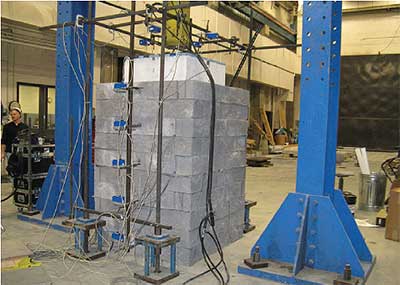 The photograph depicts a vertical column, square in cross-section, consisting of 10 tiers of rectangular masonry blocks. The column stands on the floor of a large industrial room. Vertical steel beams, painted blue, are seen to the left and right of the masonry column. A man is seen standing behind a low shelf of instruments at the left. A large concrete block connected to a cylindrical vertical shaft is pressing down on the top of the masonry column. Cables and wires are attached at various positions on the masonry column.