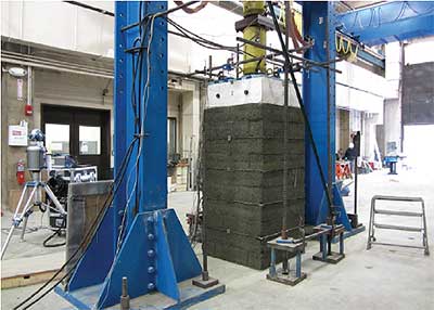 The photograph depicts a masonry-faced vertical column, square in cross-section, consisting of 10 tiers of compressed soil blocks. A thin layer of fabric is visible between every second tier of blocks. The column stands on the floor of a large industrial room. Vertical steel beams, painted blue, are seen to the left and right of the column. A large concrete block connected to a cylindrical vertical shaft is pressing down on the top of the column. Cables and wires are attached at various positions on the concrete block and vertical shaft.
