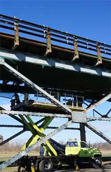 Photograph shows the side view of a multiple-span bridge, with a construction vehicle in the foreground.