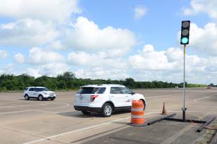 Photograph shows a roadway intersection with a traffic signal. The light is green. A car approaching the intersection from the left is stopped. Another car is moving away from the camera through the intersection.