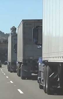 Photograph shows three tractor trailer trucks, seen from behind, traveling in the right lane of a two-lane roadway.