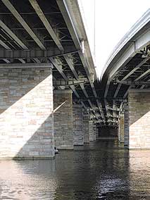 Photograph shows a view from below a highway bridge spanning a body of water. Two separate roadways merge into a single span at the center of the image
