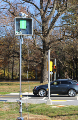 Photograph shows metal pole supporting square housing containing radar components. In the background, a car is seen passing through an intersection. Photograph shows metal pole supporting square housing containing radar components. In the background, a car is seen passing through an intersection.