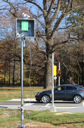 Photograph shows metal pole supporting square housing containing radar components. In the background, a car is seen passing through an intersection.