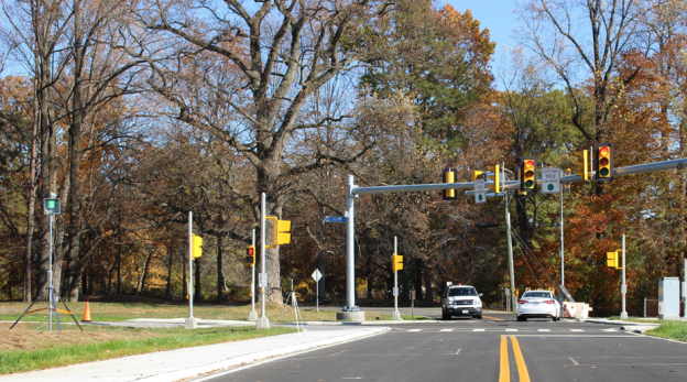 Photograph shows a four-way intersection with traffic signals. Two cars are passing through the intersection. A pole-mounted radar unit is seen on the grass adjacent to the roadway. Photograph shows a four-way intersection with traffic signals. Two cars are passing through the intersection. A pole-mounted radar unit is seen on the grass adjacent to the roadway.