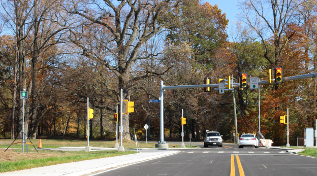 Photograph shows a four-way intersection with traffic signals. Two cars are passing through the intersection. A pole-mounted radar unit is seen on the grass adjacent to the roadway.