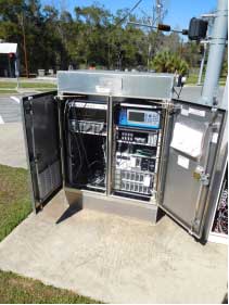 Photograph shows a metal cabinet in an outdoor setting. The inside of the cabinet has two compartments, with shelves in each compartment and electronic equipment on the shelves. Wires connect the various components of a controller that optimizes signal phase and timings for traffic signals.
