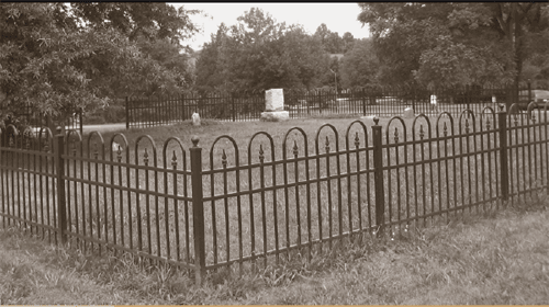 This photograph includes the entire cemetery.  One large and two small gravestones are visible.  It is surrounded by a black iron fence, and there are several trees in the background.