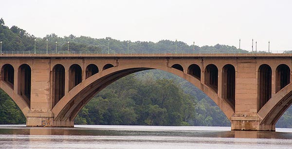 Partial side view of a large, masonry, open-spandrel, multiple-arch bridge over still water with guardrail and streetlights atop bridge. A forested hillside is in the background.