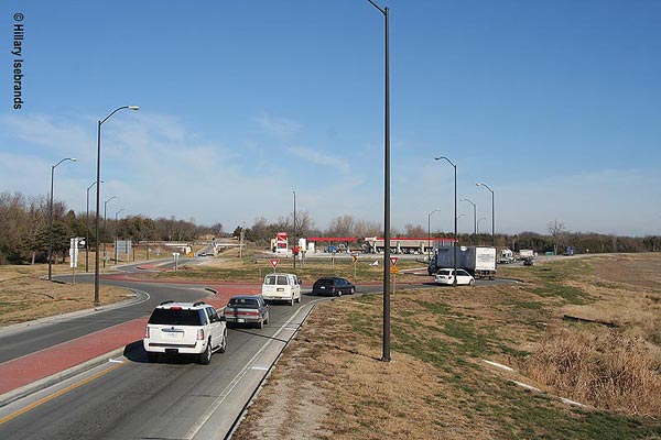 View of traffic entering a roundabout. A raised brick median separates the lanes entering and exiting the circle. Three vehicles are approaching the circle and three, including a tractor trailer truck, are moving around the right half of the circle. The center of the circle is a grassy area surrounded by a ring of brick paving. Several streetlights are around the circle and its approaches. Buildings can be seen beyond the circle.