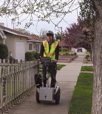 Face-on view of a man with helmet and a reflective safety vest riding a Segway®-like device on a sidewalk in a residential neighborhood. The device has handlebars and equipment mounted at the base, on the vertical pole, and at the top. Trees are to the right of the sidewalk and a picket fence is to the left of the rider. Homes and a parked pickup truck are visible in the background.
