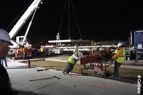 A night scene showing placement of precast concrete panels. Three workers are directing the placement of a panel suspended from a crane. Two other workers are pushing equipment on a wheeled carrier.
